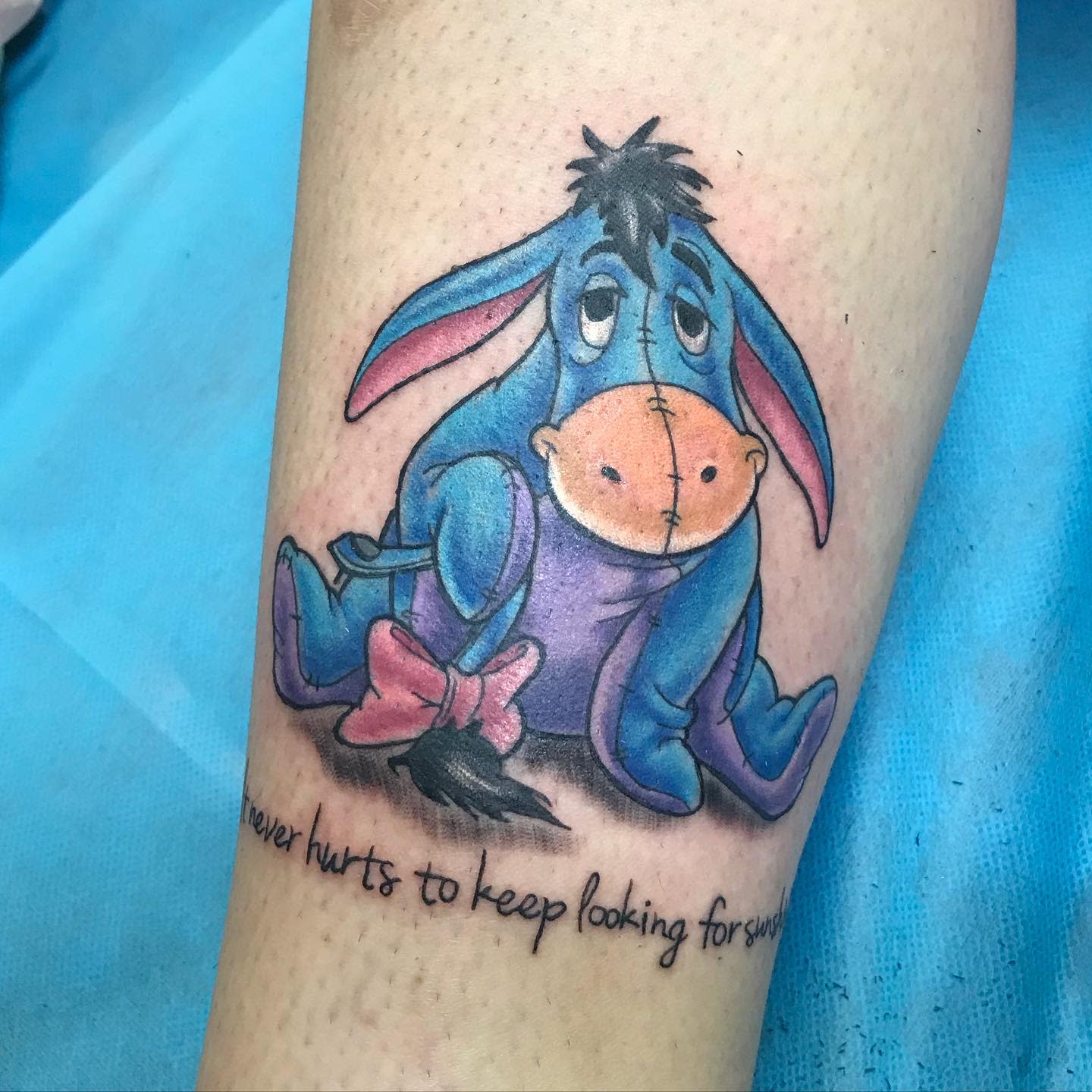 Colorful tattoo of Eeyore from Winnie the Pooh sitting with a quote beneath: 'It never hurts to keep looking for sunshine